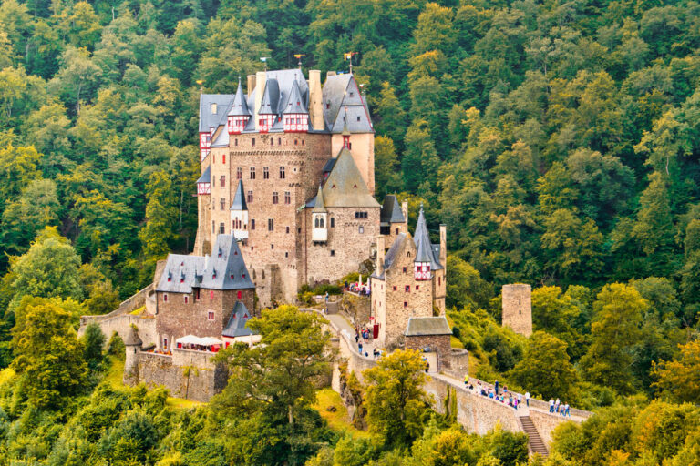 Eine Höhenburg mit Türmen, Erkern und Fachwerkgiebeln steht auf einem Felssporn mitten im dicht bewaldeten Tal der Eifel, Besucher gehen über den Aufgang zum Burgtor