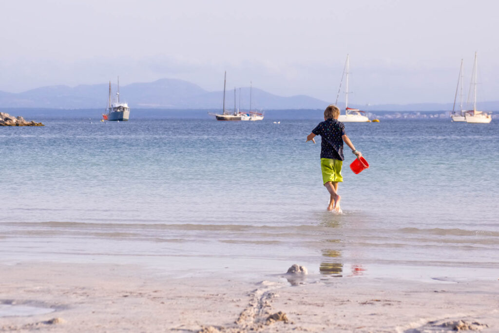 Kind steht knöcheltief weit draußen im Wasser in der Bucht von Alcúdia, dahinter ankernde Segelboote