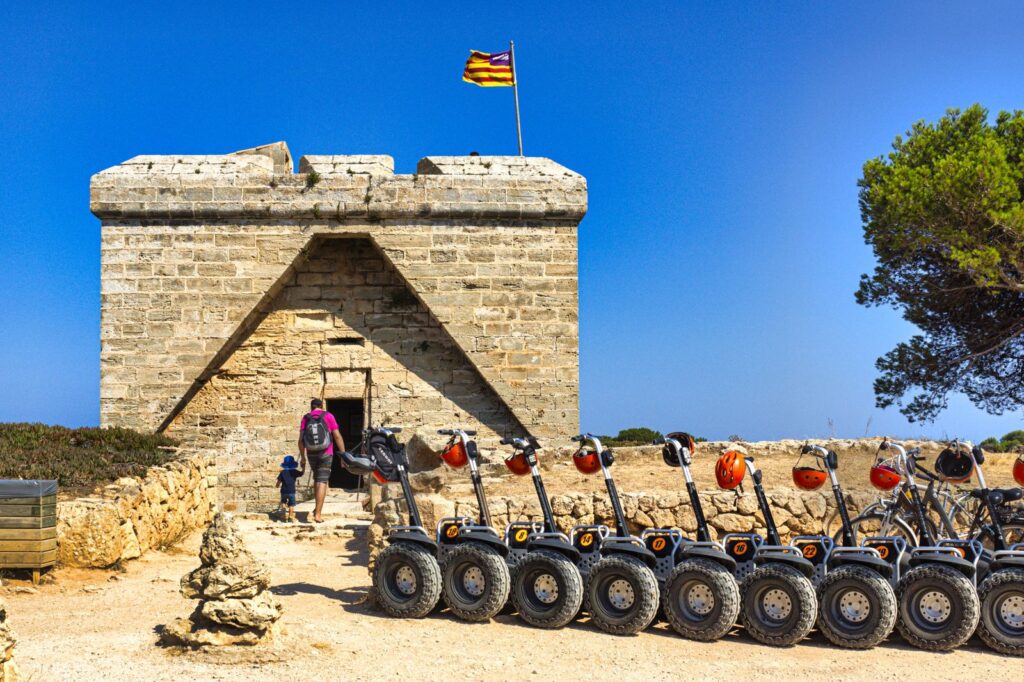 Wehrturm Castell de sa Punta de n'Amer mit mallorquinischer Flagge, davor eine Reihe geparkter Segways
