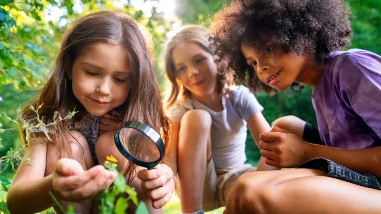 Drei Kinder hocken im Grünen, ein Mädchen betrachtet mit einer Lupe eine kleine gelbe Blüte.