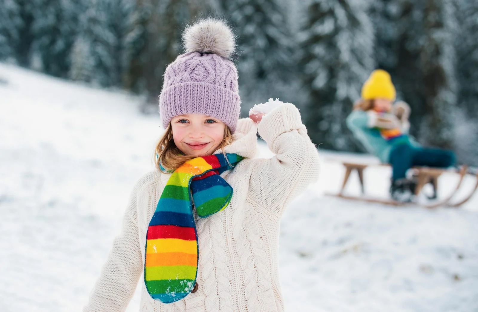 Mädchen mit Bommelmütze und Regenbogenschal wirft im verschneiten Wald einen Schneeball, im Hintergrund ein Kind auf dem Schlitten.